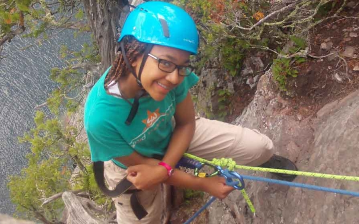 A person wearing safety gear is secured by ropes as they stand on the edge of a cliff high above a body of water and smile. 
